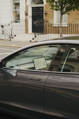 A modern car is parked on the street with a lit screen visible inside, displaying a map. The car is positioned in front of a brick building with a black door and white trim. A wrought iron fence lines the sidewalk, and a tree is present next to the car.