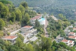 A scenic landscape features modern villas with red-tiled roofs nestled on a lush hillside surrounded by dense greenery. A winding road connects the residences, and an infinity pool can be seen near the top of the hill, overlooking the valley. The backdrop is filled with verdant trees and distant rocky hills under a clear sky.