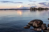 calm water with rocks nearby. In horizon two people in canoe, trees and sun peaking through clouds