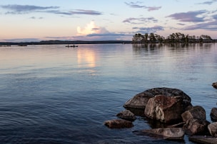calm water with rocks nearby. In horizon two people in canoe, trees and sun peaking through clouds
