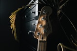 A wooden guitar headstock with tuning pegs is prominently featured. Light casts a warm glow on the instrument, highlighting the strings and the brand logo. The background is dark, with hints of plant leaves and a piece of furniture adding depth to the scene.