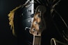 A wooden guitar headstock with tuning pegs is prominently featured. Light casts a warm glow on the instrument, highlighting the strings and the brand logo. The background is dark, with hints of plant leaves and a piece of furniture adding depth to the scene.