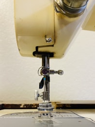 A close-up view of a sewing machine, highlighting the needle and presser foot. The image focuses on the mechanical parts with a neutral-colored background. Metal components and adjustment screws are visible, showcasing the detailed mechanics of the machine.