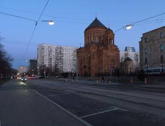 A brown brick church stands prominently with a tall, domed structure adorned with crosses on top. Surrounding the church are modern high-rise buildings. The street in the foreground is empty, with a few cars and streetlights illuminating the scene as evening approaches.
