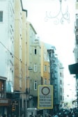 A bustling urban street scene featuring tall, narrow buildings with colorful facades ranging from mustard yellow to pale blue. There is a prominent traffic sign in the foreground that restricts vehicle access, written in Turkish. The street is filled with people, and various shop signs are visible. Overhead, decorative wrought-iron street lamps add a decorative touch, and power lines crisscross between the buildings.