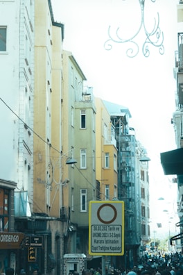 A bustling urban street scene featuring tall, narrow buildings with colorful facades ranging from mustard yellow to pale blue. There is a prominent traffic sign in the foreground that restricts vehicle access, written in Turkish. The street is filled with people, and various shop signs are visible. Overhead, decorative wrought-iron street lamps add a decorative touch, and power lines crisscross between the buildings.