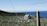 A scenic view of a large cemetery surrounded by a chain-link fence. Numerous tombstones are scattered across the green grass, and a small chapel or building stands in the center. In the background, a clear blue sky meets the deep blue sea, creating a serene atmosphere.