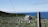 A scenic view of a large cemetery surrounded by a chain-link fence. Numerous tombstones are scattered across the green grass, and a small chapel or building stands in the center. In the background, a clear blue sky meets the deep blue sea, creating a serene atmosphere.
