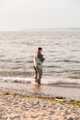 A person in waders stands at the edge of the sea with a fishing rod, casting into the waves. The shoreline is speckled with small rocks and patches of seaweed, and the water reflects the soft light of either early morning or late afternoon.