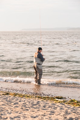 A person in waders stands at the edge of the sea with a fishing rod, casting into the waves. The shoreline is speckled with small rocks and patches of seaweed, and the water reflects the soft light of either early morning or late afternoon.