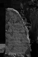 An ancient stone with inscriptions carved in a script resembling Greek, standing upright in a possibly archaeological or historical site. The stone has an uneven surface with weathered edges, surrounded by a rocky and uneven terrain.