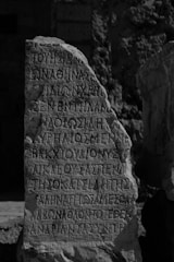 An ancient stone with inscriptions carved in a script resembling Greek, standing upright in a possibly archaeological or historical site. The stone has an uneven surface with weathered edges, surrounded by a rocky and uneven terrain.