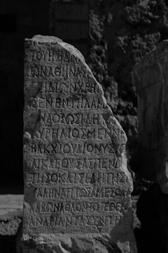 An ancient stone with inscriptions carved in a script resembling Greek, standing upright in a possibly archaeological or historical site. The stone has an uneven surface with weathered edges, surrounded by a rocky and uneven terrain.