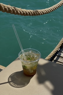 A plastic cup filled with a refreshing drink, garnished with mint leaves and ice, is placed on a light-colored surface. In the background, turquoise water and a thick rope create a nautical atmosphere, illuminated by sunlight casting shadows.