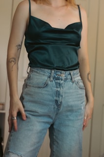 A person is wearing a dark green satin camisole and high-waisted light blue jeans. The camisole features thin straps, while the jeans have a casual, worn look with slight distressing. Tattoos are visible on the person's arms, and they are wearing rings on multiple fingers.