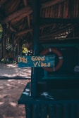 A rustic wooden sign displaying the phrase 'Only Good Vibes' in yellow letters is attached to a dark blue painted post under a thatched roof. The background includes a life preserver and a sandy area with scattered sunlight filtering through trees.