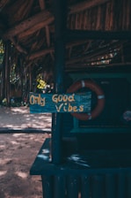 A rustic wooden sign displaying the phrase 'Only Good Vibes' in yellow letters is attached to a dark blue painted post under a thatched roof. The background includes a life preserver and a sandy area with scattered sunlight filtering through trees.