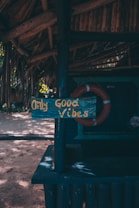 A rustic wooden sign displaying the phrase 'Only Good Vibes' in yellow letters is attached to a dark blue painted post under a thatched roof. The background includes a life preserver and a sandy area with scattered sunlight filtering through trees.