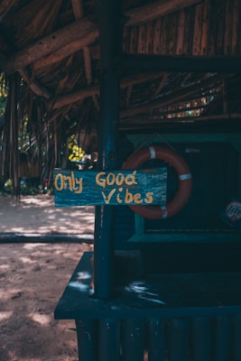 A rustic wooden sign displaying the phrase 'Only Good Vibes' in yellow letters is attached to a dark blue painted post under a thatched roof. The background includes a life preserver and a sandy area with scattered sunlight filtering through trees.