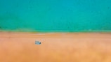 Aerial view of a tranquil beach scene with a clear turquoise sea meeting a golden sandy shore. A set of blue lounge chairs is neatly arranged on the sand, with a single person lying on one of them.