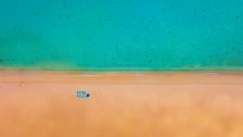 Aerial view of a tranquil beach scene with a clear turquoise sea meeting a golden sandy shore. A set of blue lounge chairs is neatly arranged on the sand, with a single person lying on one of them.