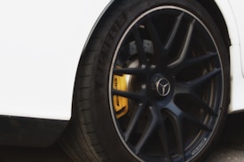 A close-up of a car wheel featuring a black alloy rim with a visible disc brake system and a yellow brake caliper. The tire is branded with 'Michelin' and the car appears to have a white body. The center of the alloy wheel has a car logo.