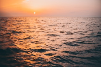 A calm ocean scene at sunset with gentle waves reflecting soft orange and blue hues.