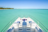 A yacht's deck extends towards a vast, calm turquoise ocean with a clear blue sky overhead. Towels and gear are scattered across the deck, and shadows suggest the presence of a person standing. A tropical coastline with palm trees is visible in the background.