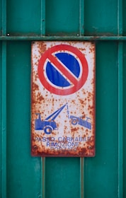 A rusty sign depicting a no parking symbol with a red circle and a blue background, alongside icons of two tow trucks. The text reads 'PASSO CARRABILE RIMOZIONE FORZATA'. The sign is mounted on a textured, green metal surface with visible corrosion.