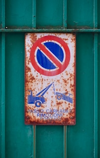 A rusty sign depicting a no parking symbol with a red circle and a blue background, alongside icons of two tow trucks. The text reads 'PASSO CARRABILE RIMOZIONE FORZATA'. The sign is mounted on a textured, green metal surface with visible corrosion.