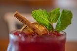 A close-up of a refreshing beverage garnished with fresh mint leaves, a slice of dried lime, and a stick of cinnamon. The drink has a rich burgundy color, and the glass is filled with crushed ice. The background is blurred, putting emphasis on the drink.