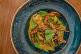 A beautifully plated dish of seafood consisting of crab legs and shrimp, garnished with fresh basil leaves. The ingredients are placed on a bed of creamy sauce, within a deep blue ceramic dish, set against a wooden table backdrop.