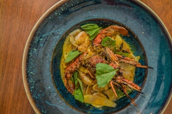A beautifully plated dish of seafood consisting of crab legs and shrimp, garnished with fresh basil leaves. The ingredients are placed on a bed of creamy sauce, within a deep blue ceramic dish, set against a wooden table backdrop.