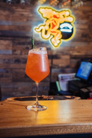 A vibrant orange cocktail served in a tall glass on a wooden bar counter. The cocktail is garnished with a sprig of rosemary. In the background, there is a blurred neon sign depicting a stylized figure, adding a lively touch to the ambiance.