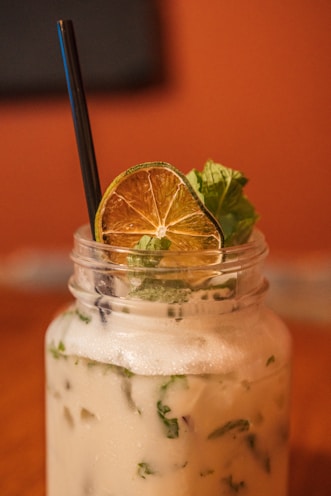 A creamy drink is served in a mason jar, topped with a slice of dried lime and fresh mint leaves. A black straw sticks out from the jar. The background is a soft, warm tone, possibly a wooden table surface and an out-of-focus wall.