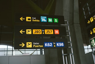 Several airport signs indoors provide directions to various exits and gates. The signs include yellow, black, green, and blue colors with white directional arrows. The text is in both Spanish and English, with labels for different terminal sections and gate ranges.