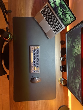 A wooden desk setup includes a laptop with a leaf-themed wallpaper, a mechanical keyboard, a gaming mouse, and a large monitor displaying the same wallpaper. Items such as a microphone, headphones, and a few mugs are also present. The lighting is warm, emanating from beneath the monitor and the desk's edge.