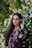 A woman with long dark hair is standing against a backdrop of lush greenery and white flowers. She is wearing a floral-patterned outfit with shades of purple and black. Her right hand is gently touching a white flower near her face, and she is gazing warmly at the camera.
