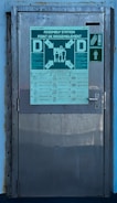 A metal door with signs and instructions displayed on it. The largest sign at the top reads 'Assembly Station' and includes various arrows and figures. Below this are detailed instructions with visual icons corresponding to different safety procedures. There is a smaller green sign to the right depicting a staircase and a figure, indicating an emergency exit or path.