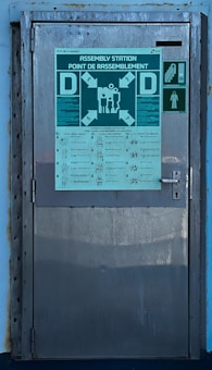 A metal door with signs and instructions displayed on it. The largest sign at the top reads 'Assembly Station' and includes various arrows and figures. Below this are detailed instructions with visual icons corresponding to different safety procedures. There is a smaller green sign to the right depicting a staircase and a figure, indicating an emergency exit or path.