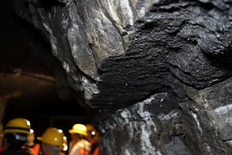 A rocky underground environment, likely a cave or mine, with layered mineral formations visible on the walls. Several individuals wearing yellow hard hats and safety vests can be seen, indicating a guided tour or exploration activity.