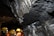 A rocky underground environment, likely a cave or mine, with layered mineral formations visible on the walls. Several individuals wearing yellow hard hats and safety vests can be seen, indicating a guided tour or exploration activity.