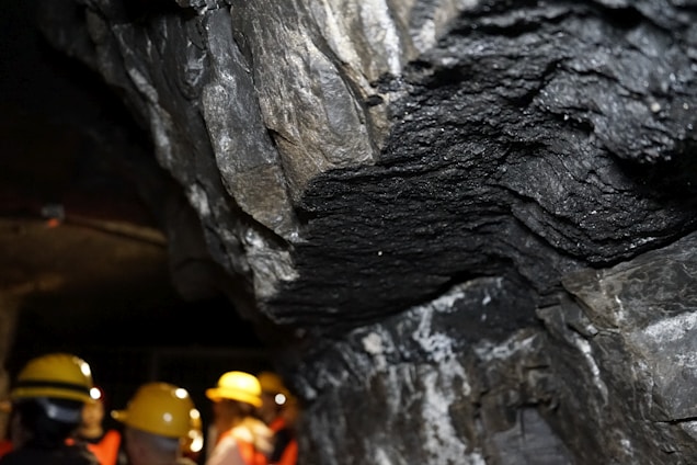 A rocky underground environment, likely a cave or mine, with layered mineral formations visible on the walls. Several individuals wearing yellow hard hats and safety vests can be seen, indicating a guided tour or exploration activity.