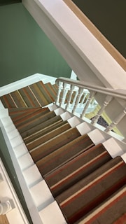 A staircase with white railings and a carpet featuring green, brown, and red stripes. The walls are painted green, and the stairs ascend in a geometric pattern.