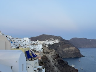 Whitewashed buildings with blue domes perched on a steep hillside overlook the serene blue waters of a bay. Several boats and yachts are moored in the calm waters below. The scene captures a tranquil coastal village setting at dusk, with hillside terrain extending into the distance.