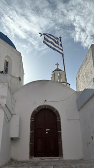 A traditional white building features a dark wooden arched door in the center. Above the building is a Greek flag flying, with a blue and white striped design. There is also a small belfry with a cross on top. The sky is partially cloudy with sunlight illuminating the scene.