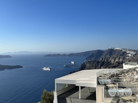 A scenic view of a coastal area with several cruise ships sailing on the blue sea, bordered by rugged hills. In the foreground, there's a modern terrace with empty tables and chairs, overlooking the water. The clear blue sky adds a serene atmosphere to the scene.