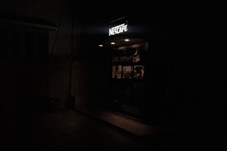 A dimly lit small coffee shop with a sign reading 'Nescafe'. Inside, a person is visible behind the counter, likely preparing or serving drinks. The interior is faintly illuminated, creating a cozy atmosphere amidst the surrounding darkness.