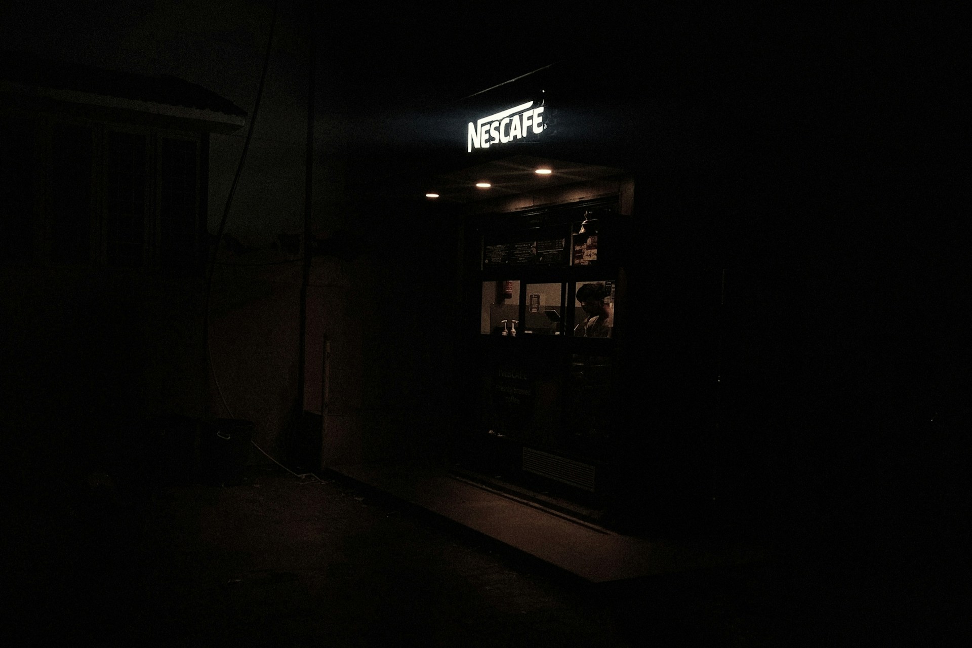 A dimly lit small coffee shop with a sign reading 'Nescafe'. Inside, a person is visible behind the counter, likely preparing or serving drinks. The interior is faintly illuminated, creating a cozy atmosphere amidst the surrounding darkness.