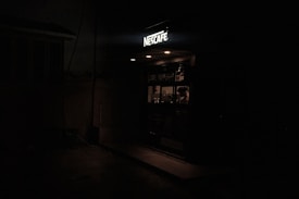 A dimly lit small coffee shop with a sign reading 'Nescafe'. Inside, a person is visible behind the counter, likely preparing or serving drinks. The interior is faintly illuminated, creating a cozy atmosphere amidst the surrounding darkness.
